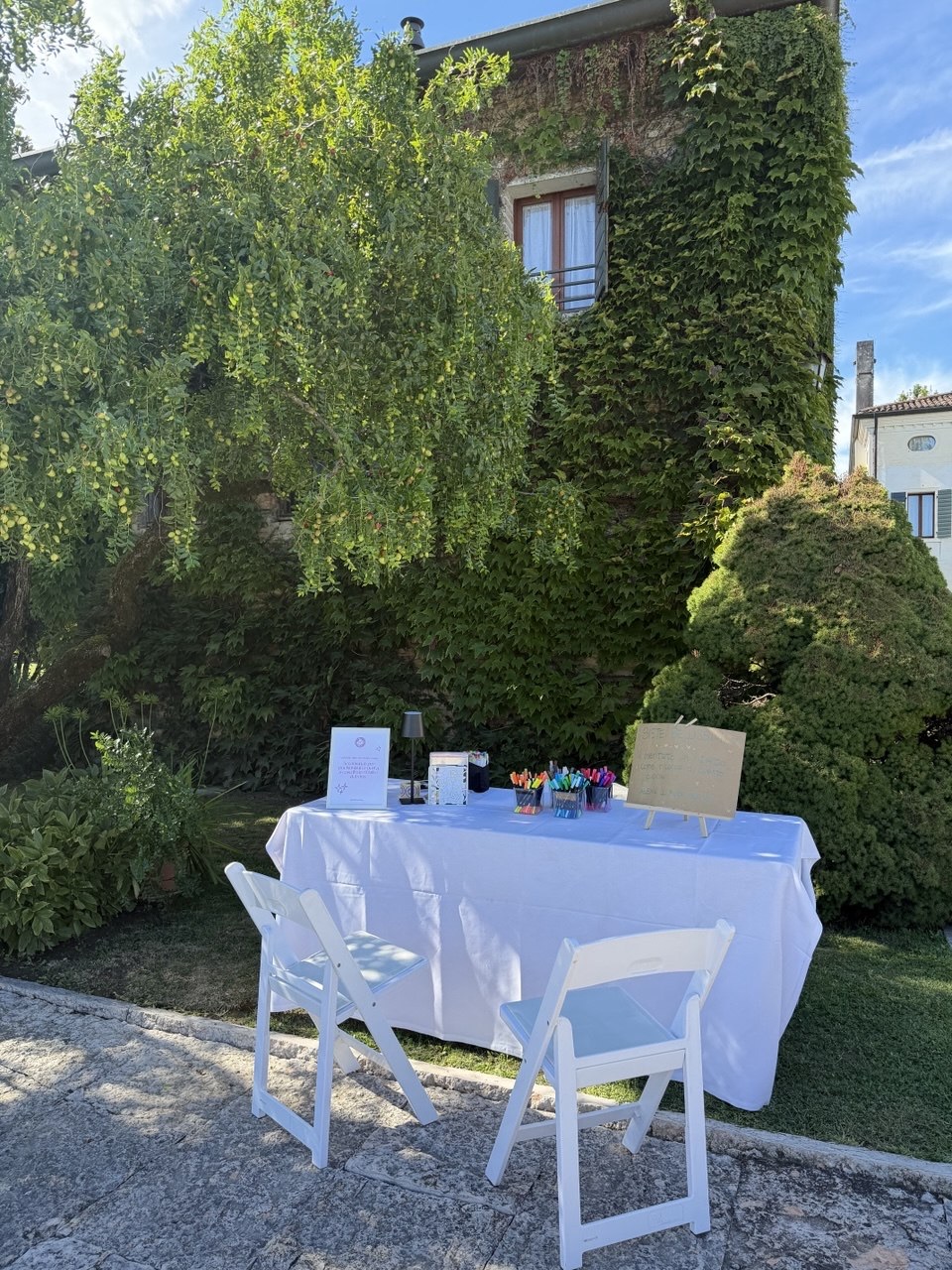 La postazione allestita in giardino: tavolo bianco con i pennarelli e il cartello, due sedie per gli ospiti, davanti a una parete di edera